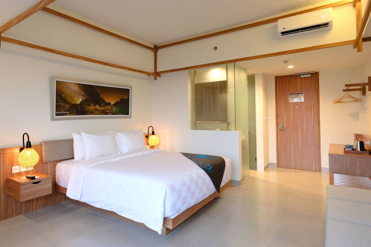 Een hotelkamer met een bed, nachtkastjes, een bureau, een stoel, een spiegel, een deur en een airconditioning.