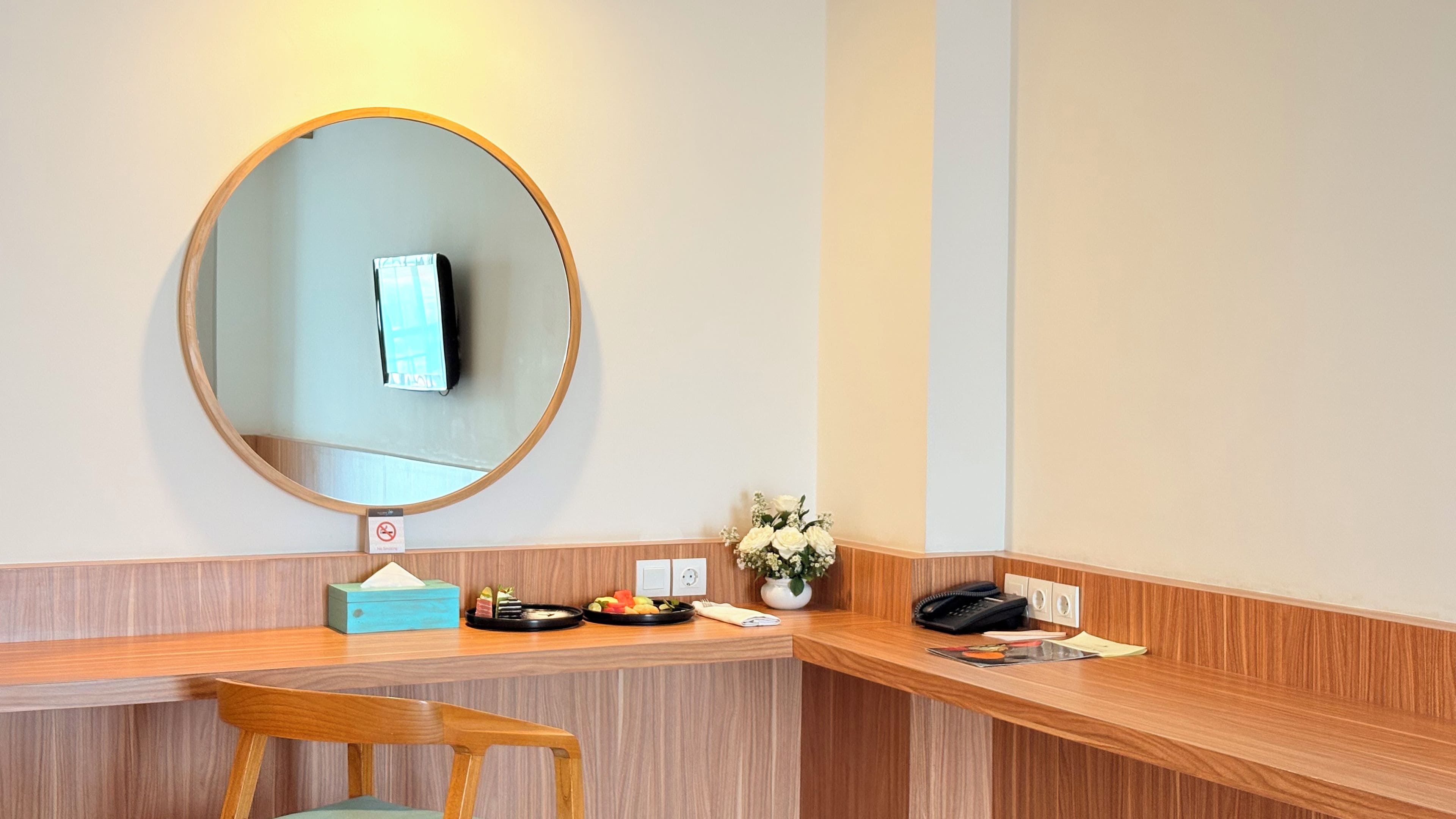 A hotel room with a wooden desk, a mirror, a vase with flowers, a telephone, and a small table with a fruit plate.