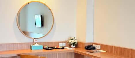 A hotel room with a wooden desk, a mirror, a vase with flowers, a telephone, and a small table with a fruit plate.