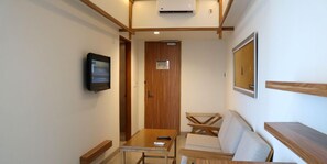 Un couloir étroit avec une table en bois et un canapé, un téléviseur fixé au mur et une porte avec une pancarte.