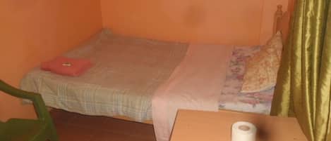 A small room with a single bed, a wooden table with a roll of toilet paper, and a green chair.