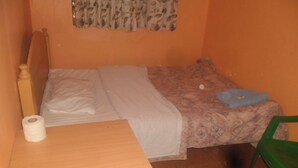 A small, single-bed room with a wooden headboard, a green plastic chair, and a small table with a roll of toilet paper. - Magi Guest House (Thika)