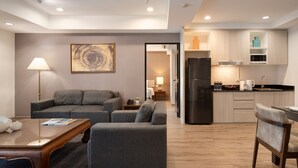 A modern living room with a grey sofa, a wooden coffee table, and a dining area with a chair. A kitchen with a black refrigerator and wooden cabinets is visible in the background. - Aspira Residence Ruamrudee (Bangkok)
