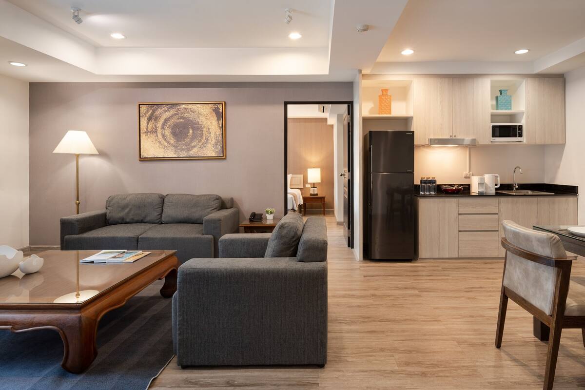 A modern living room with a grey sofa, a wooden coffee table, and a dining area with a chair. A kitchen with a black refrigerator and wooden cabinets is visible in the background.