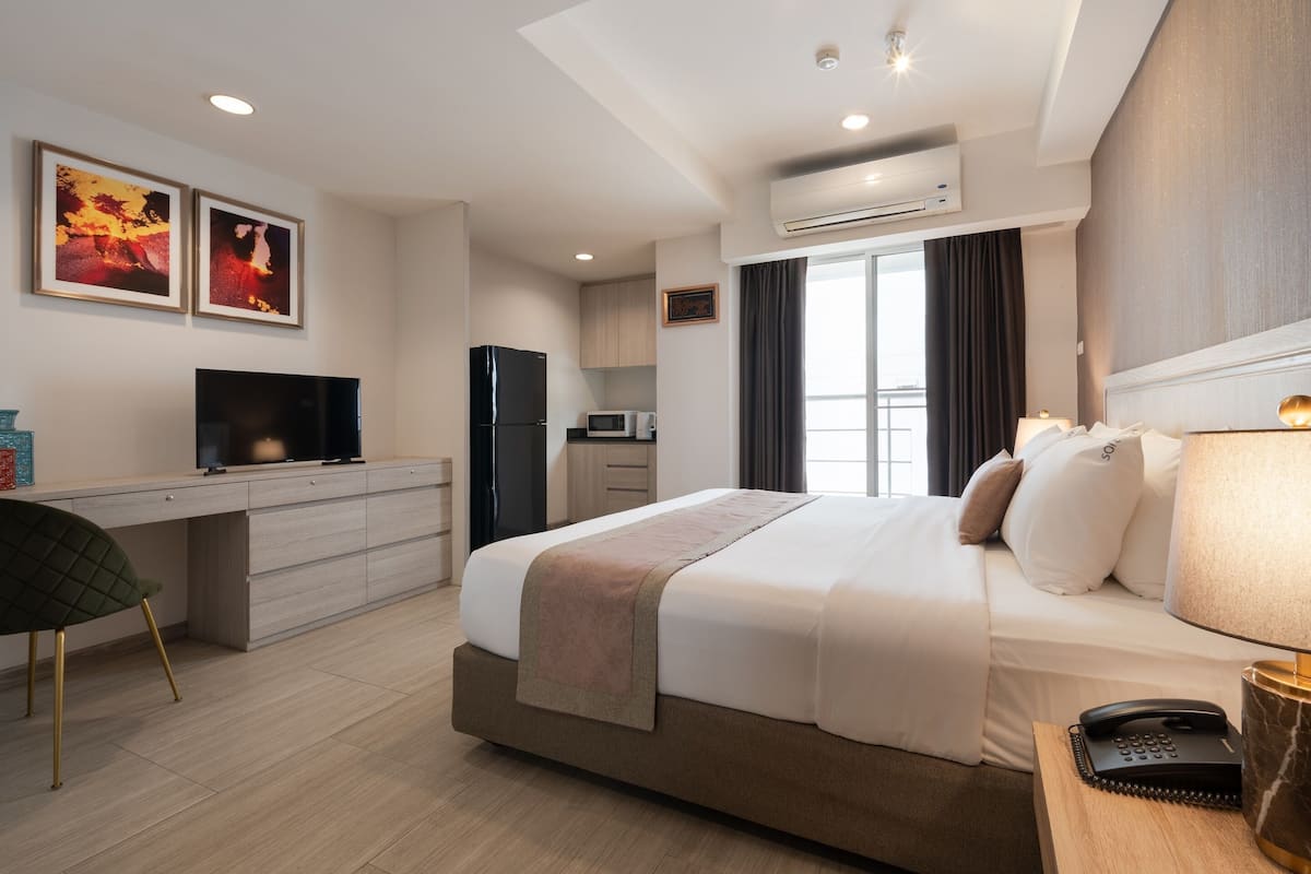 A modern hotel room with a bed, desk, TV, kitchenette, and artwork on the wall.