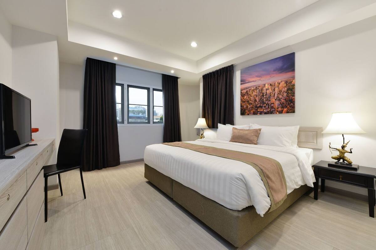 A hotel room with a large bed, a desk with a TV, a chair, two bedside tables with lamps, and a cityscape painting on the wall.