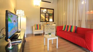 A living room with a red sofa, a white armchair, a small white table, a TV on a stand, and a window with sheer curtains.