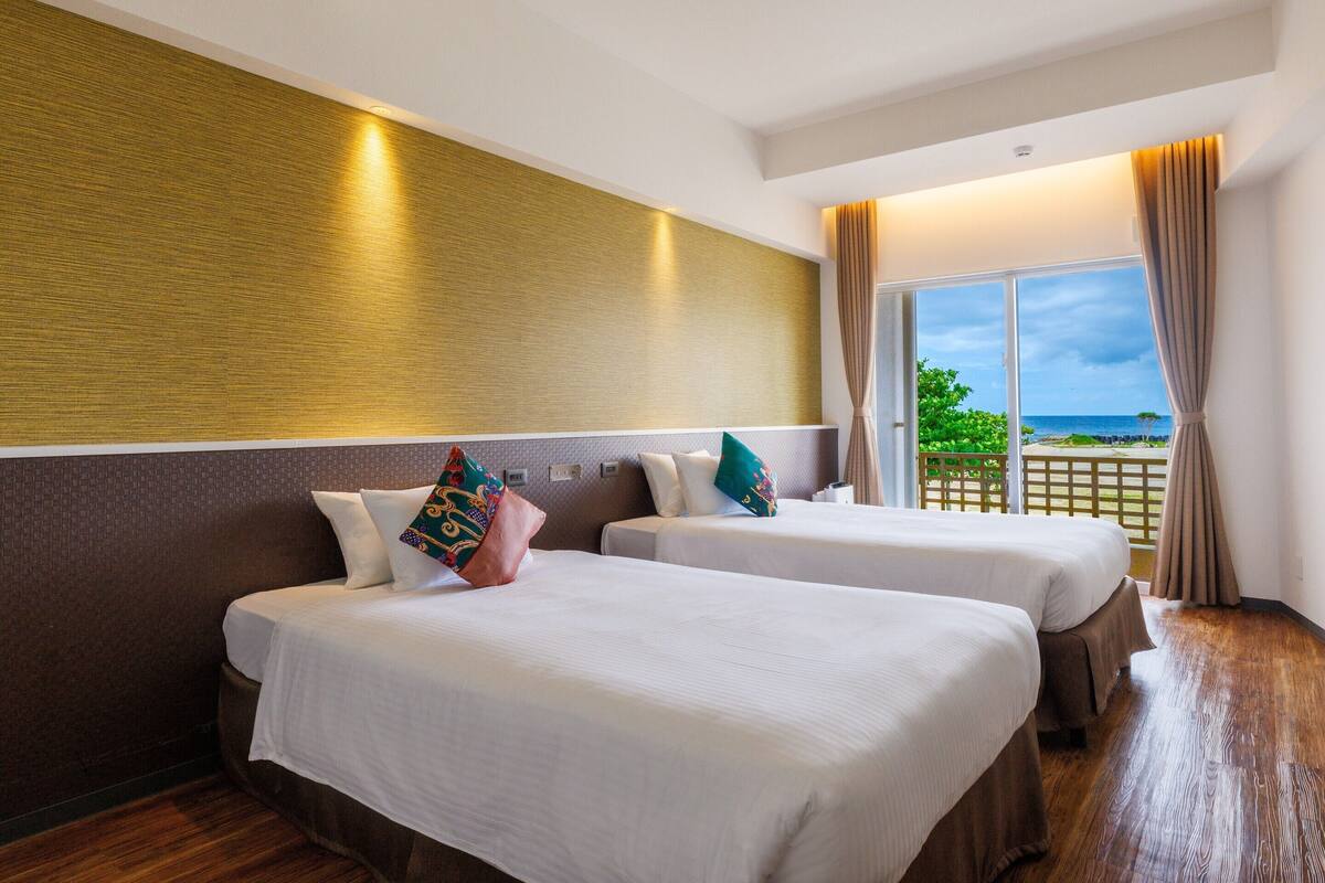 A hotel room with two beds, a large window with a view of the sea, and a wooden floor.