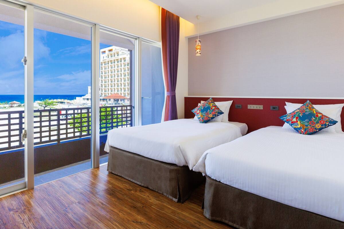 A hotel room with two beds, a balcony with a view of the ocean and a building, and a red wall.