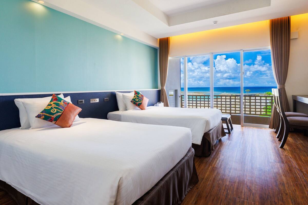 A hotel room with two beds, a balcony with a view of the ocean, and a wooden floor.