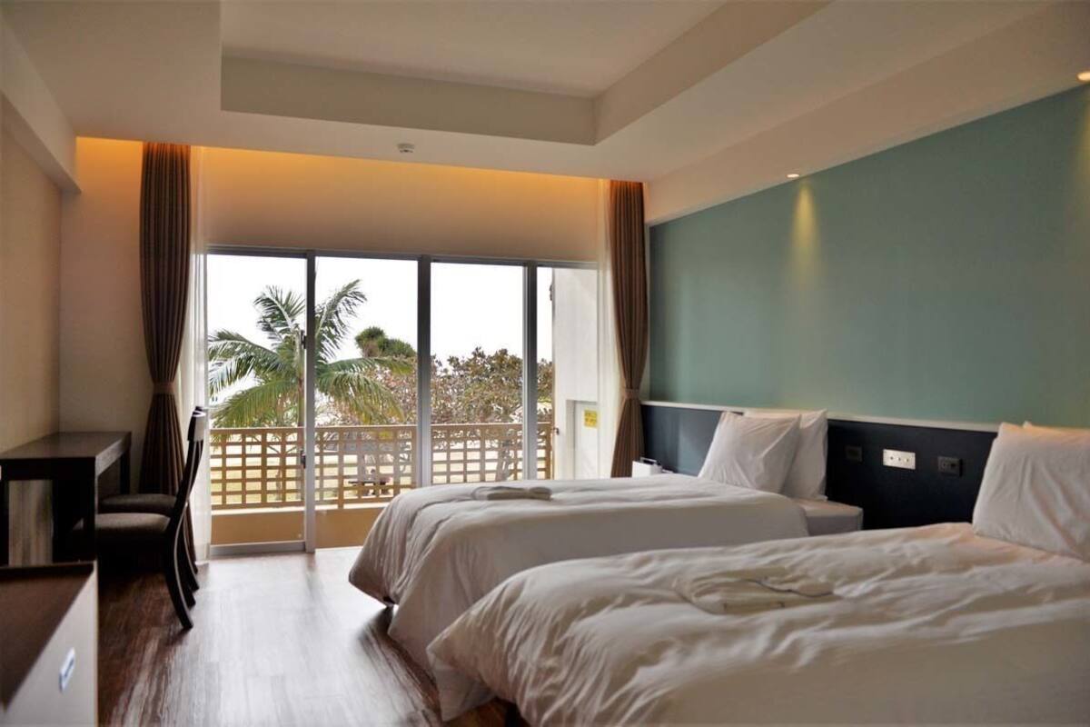 A hotel room with two beds, a desk, and a view of palm trees through a large window.