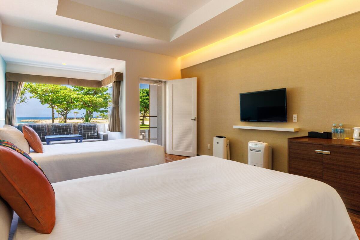 A hotel room with two beds, a TV, a desk, and a view of the beach.