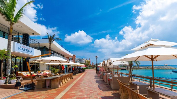 Un paseo marítimo con terrazas al aire libre, palmeras y vista al mar.