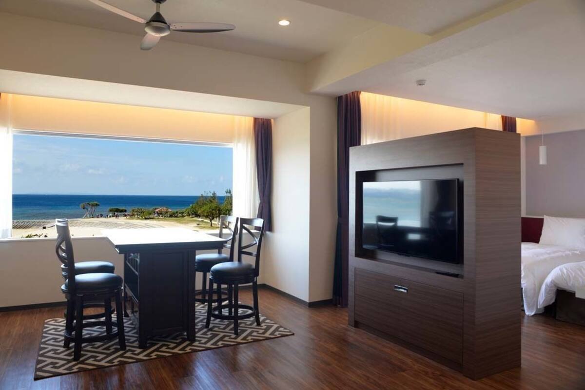A hotel room with a TV, a dining area with chairs, a ceiling fan, and a view of the beach.