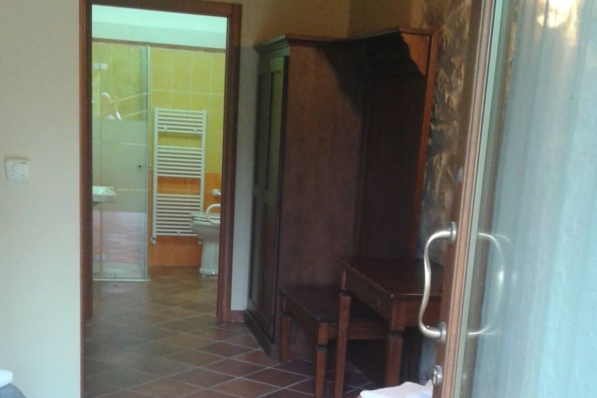 Una habitación con piso de baldosas, techo de madera, cama con cubrecama floral, aparador de madera y puerta de vidrio.