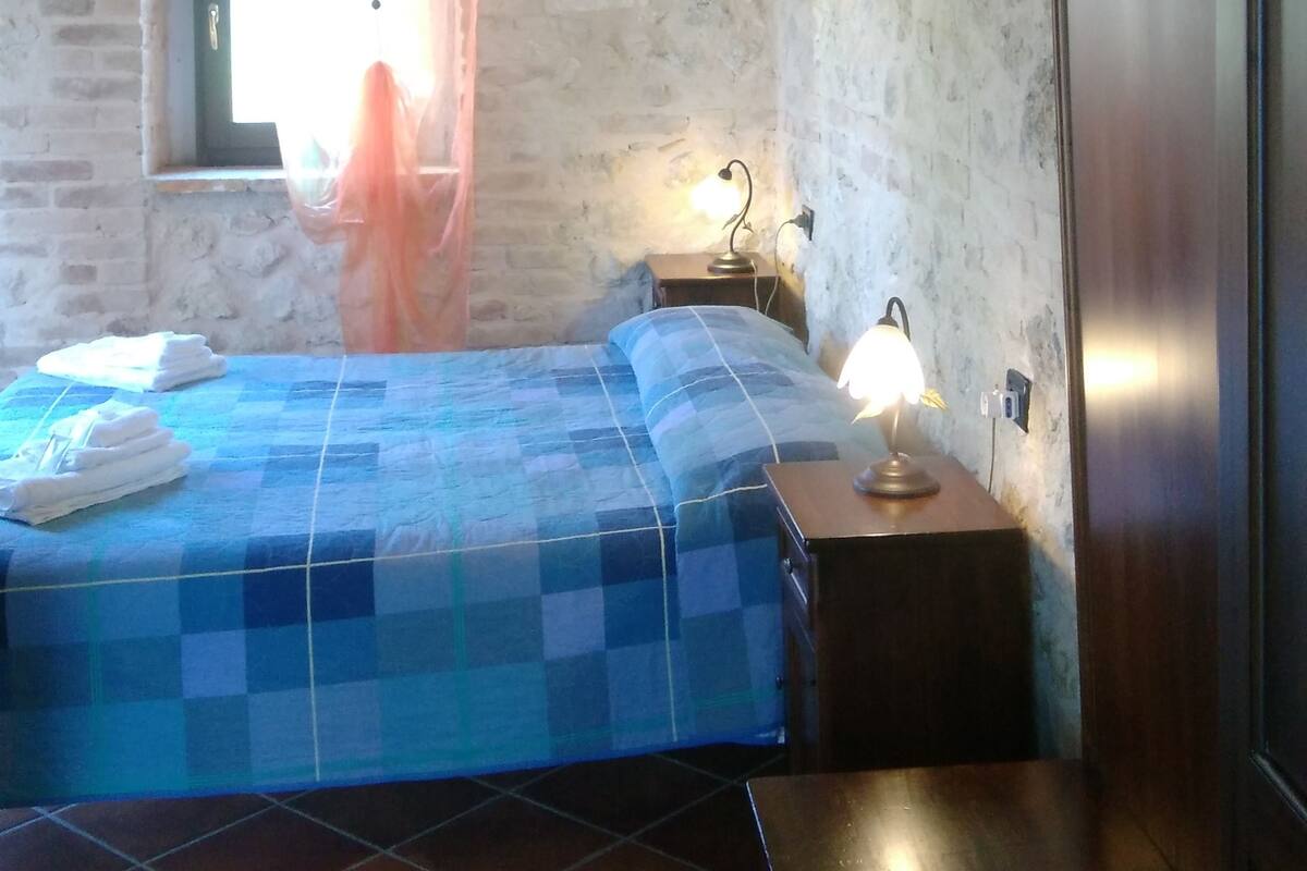 Un dormitorio con edredón a cuadros, techo de madera, pared de piedra, ventana con cortina y una lámpara de noche.
