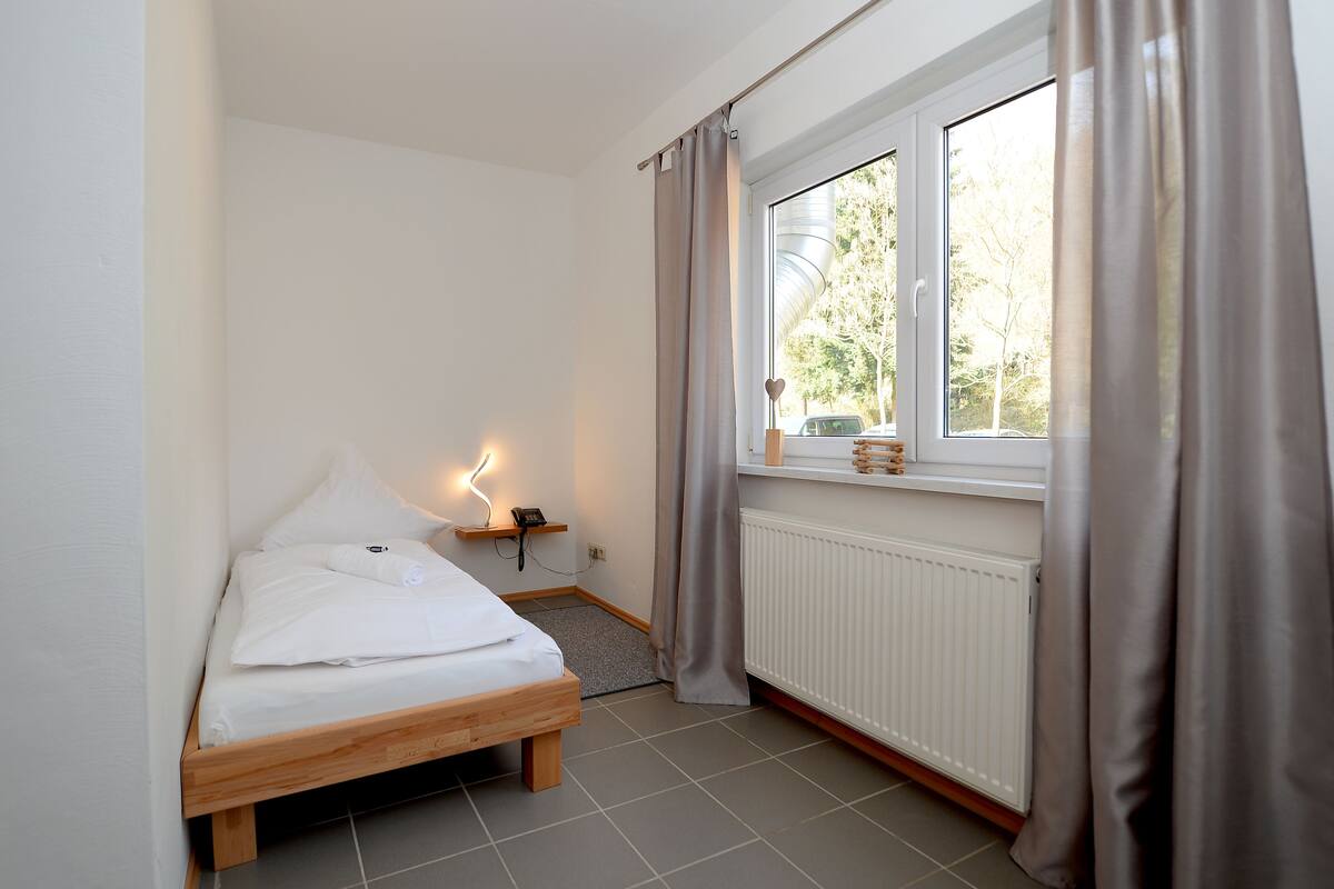 A small, neatly arranged bedroom with a single bed, a bedside table with a lamp, a window with curtains, and a radiator.