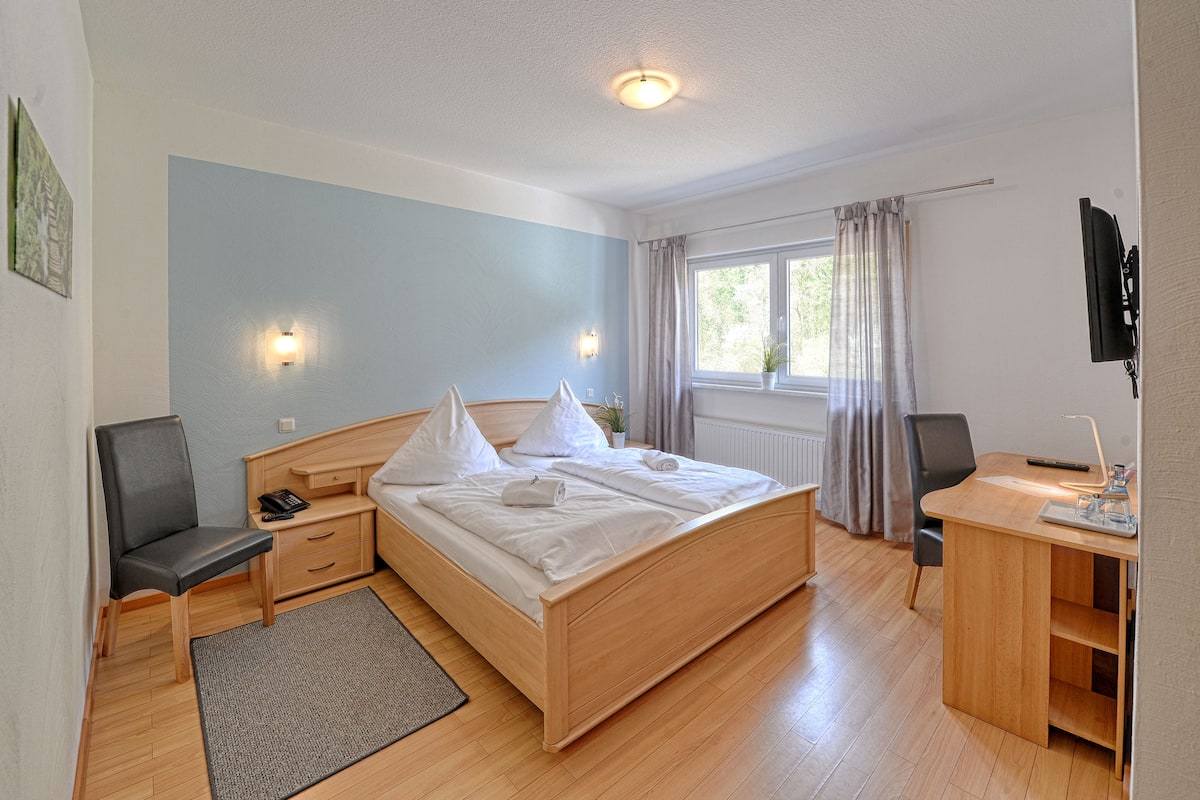 A bedroom with a bed, bedside tables, a desk, a chair, a window with curtains, and a wall-mounted TV.