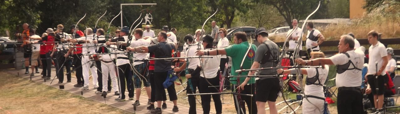 A group of people in white shirts and black pants, some holding bows, standing in a line outdoors.