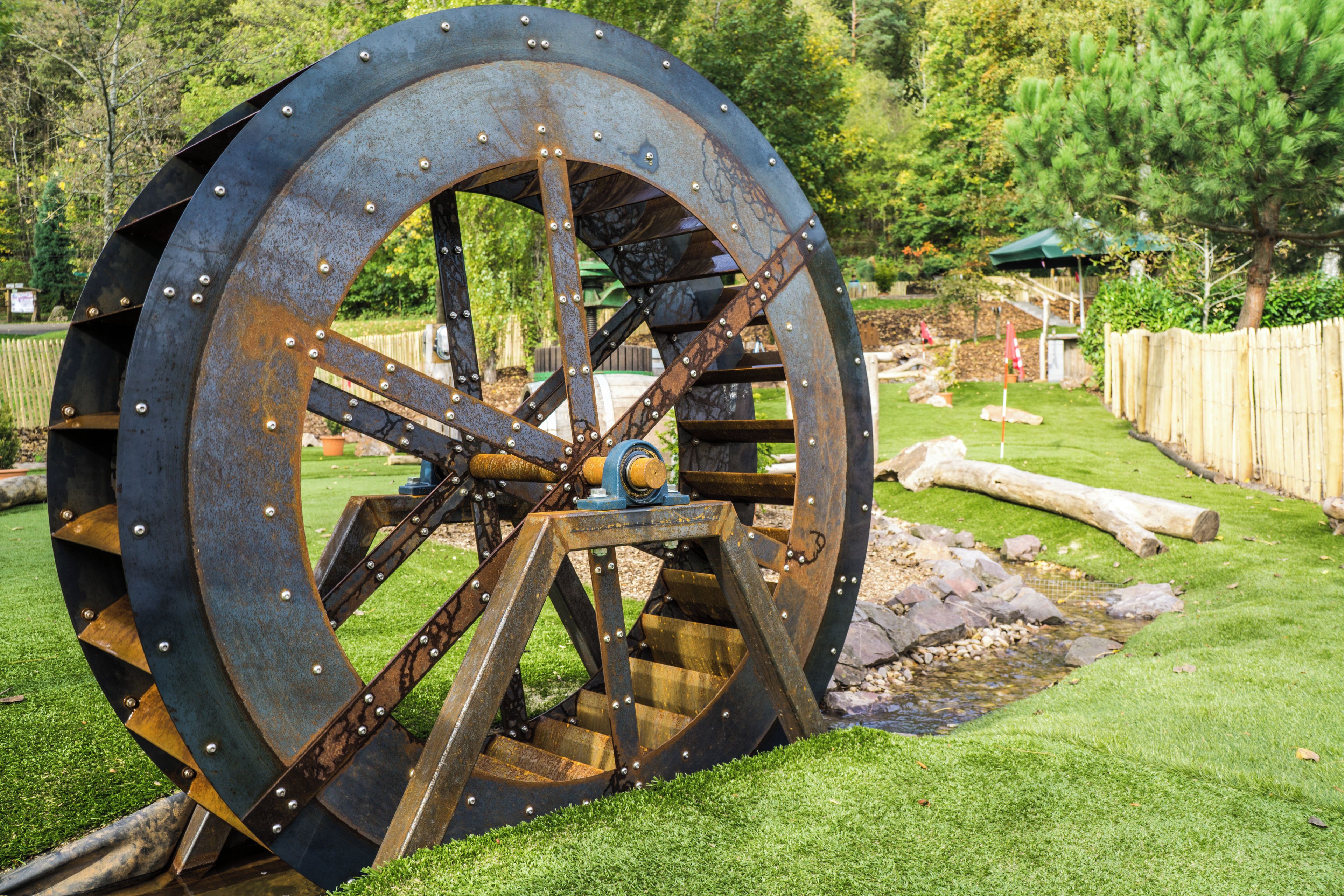 A large, rusted water wheel with a metal structure and gears, set in a grassy area with trees and a wooden fence in the background.