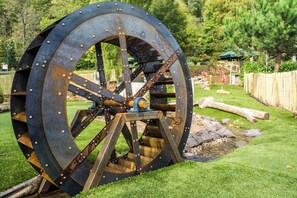 A large, rusted water wheel with a metal structure and gears, set in a grassy area with trees and a wooden fence in the background. - Hotel Restaurant Litermont (Nalbach)