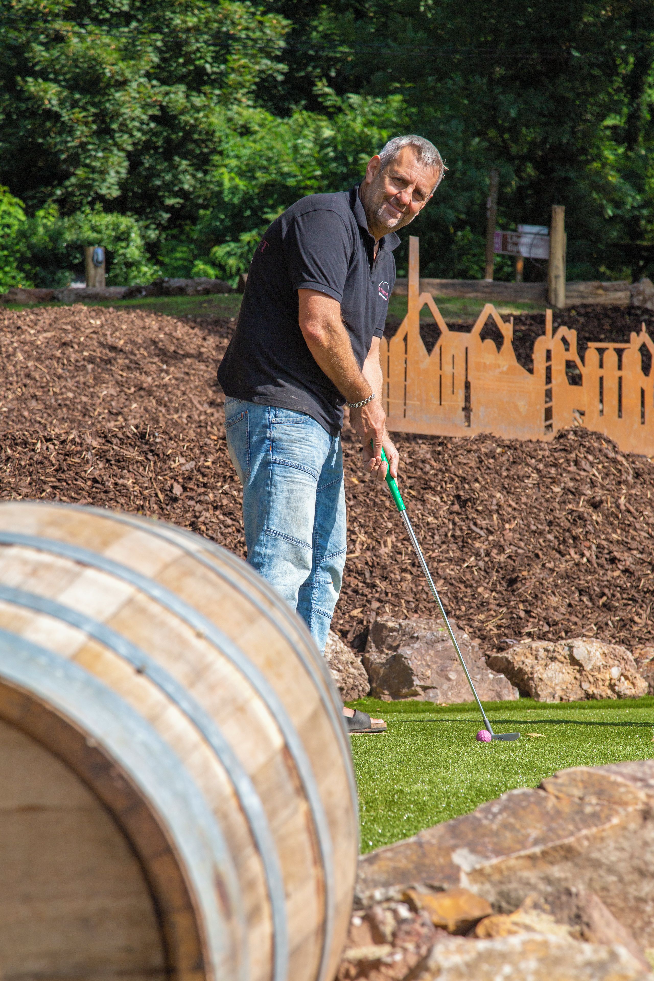 Eine Person spielt Golf auf einem Golfplatz mit einem Holzzaun und Bäumen im Hintergrund.