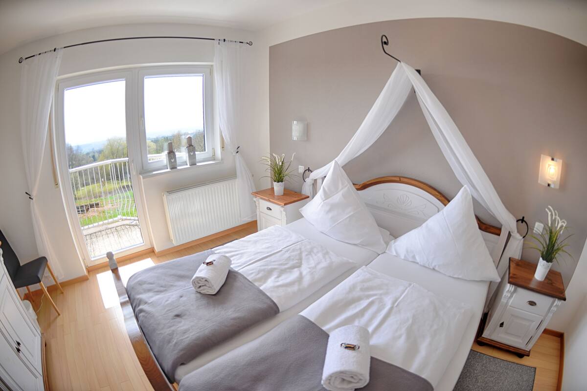 A bedroom with a double bed, a canopy bed, a window with curtains, a bedside table with a plant, and a view of the outdoors.