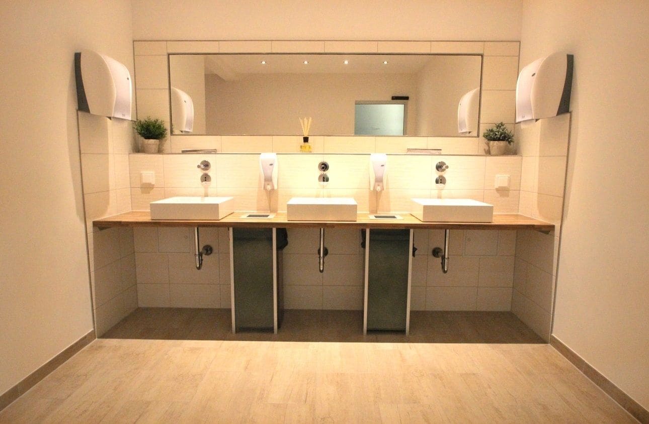Un baño moderno con tres lavamanos, un espejo grande y secamanos automáticos.