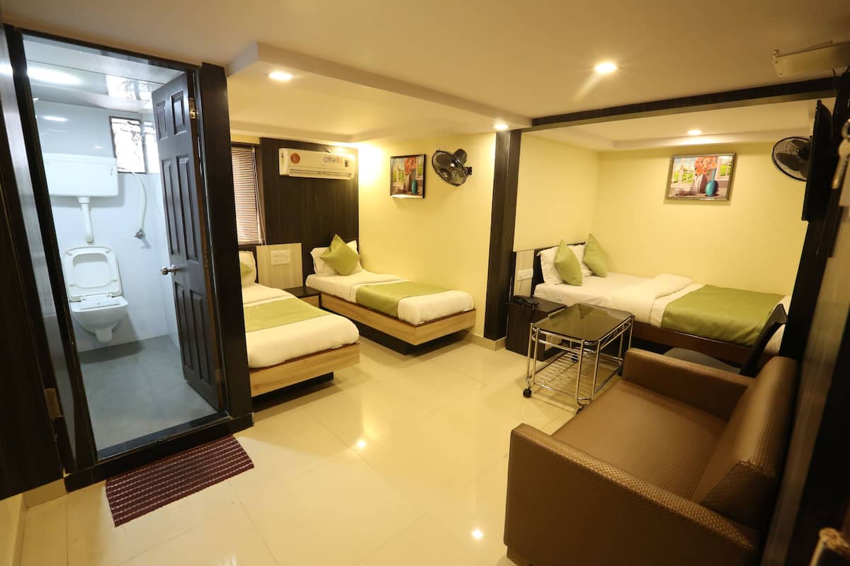 Quarto de hotel com duas camas de solteiro, um sofá, uma mesinha, um ventilador, um sanitário e uma janela.