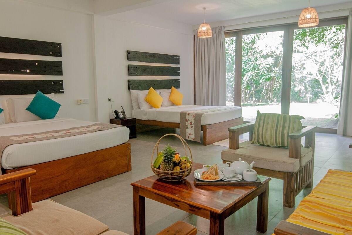 A hotel room with two beds, a sofa, a dining table with breakfast items, and large windows with a view of greenery.