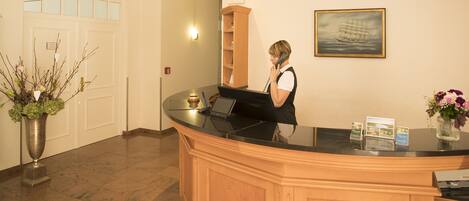 A reception desk with a receptionist, a potted plant, a framed picture on the wall, and a door in the background.
