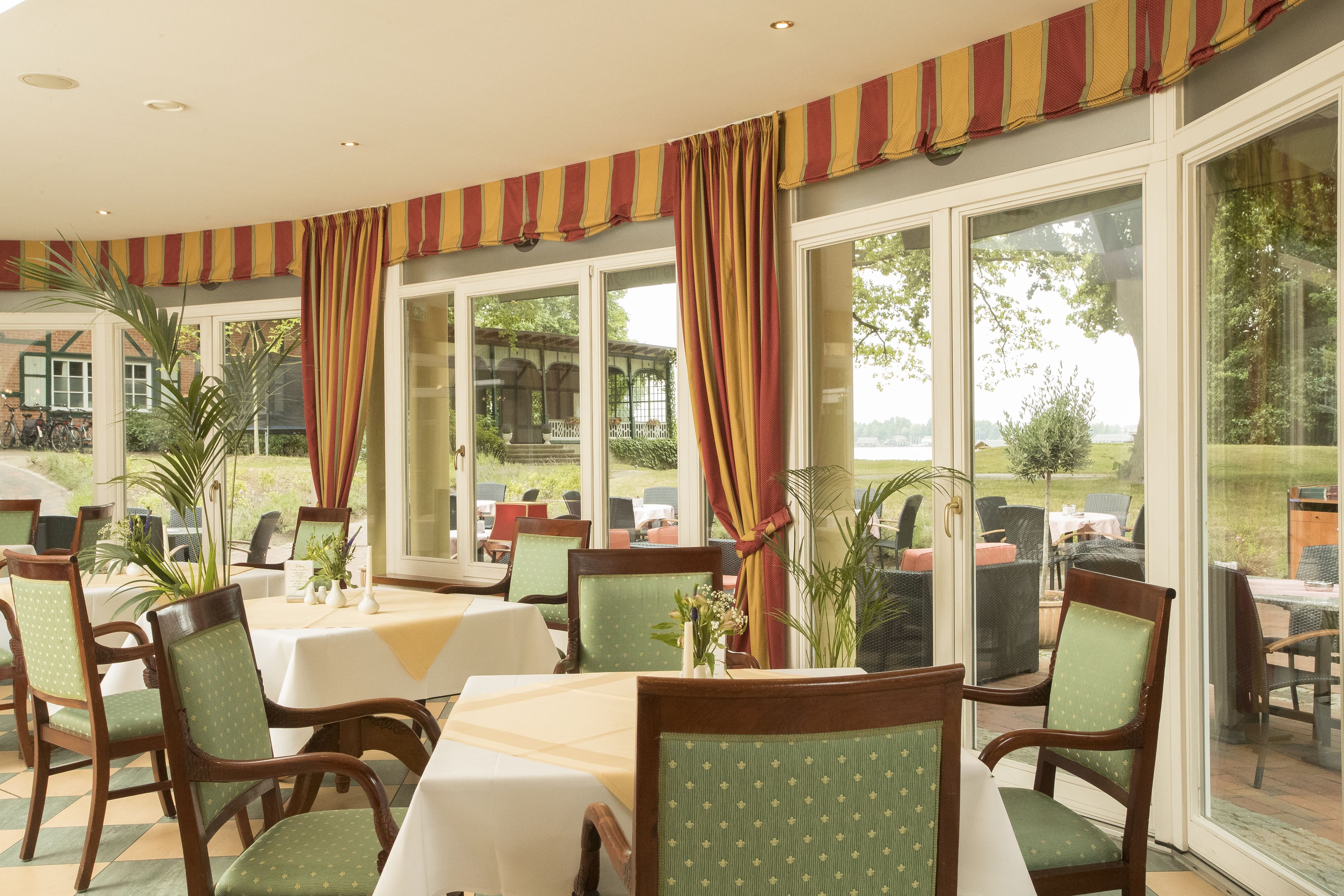 A dining area with tables and chairs, large windows with curtains, and outdoor views.