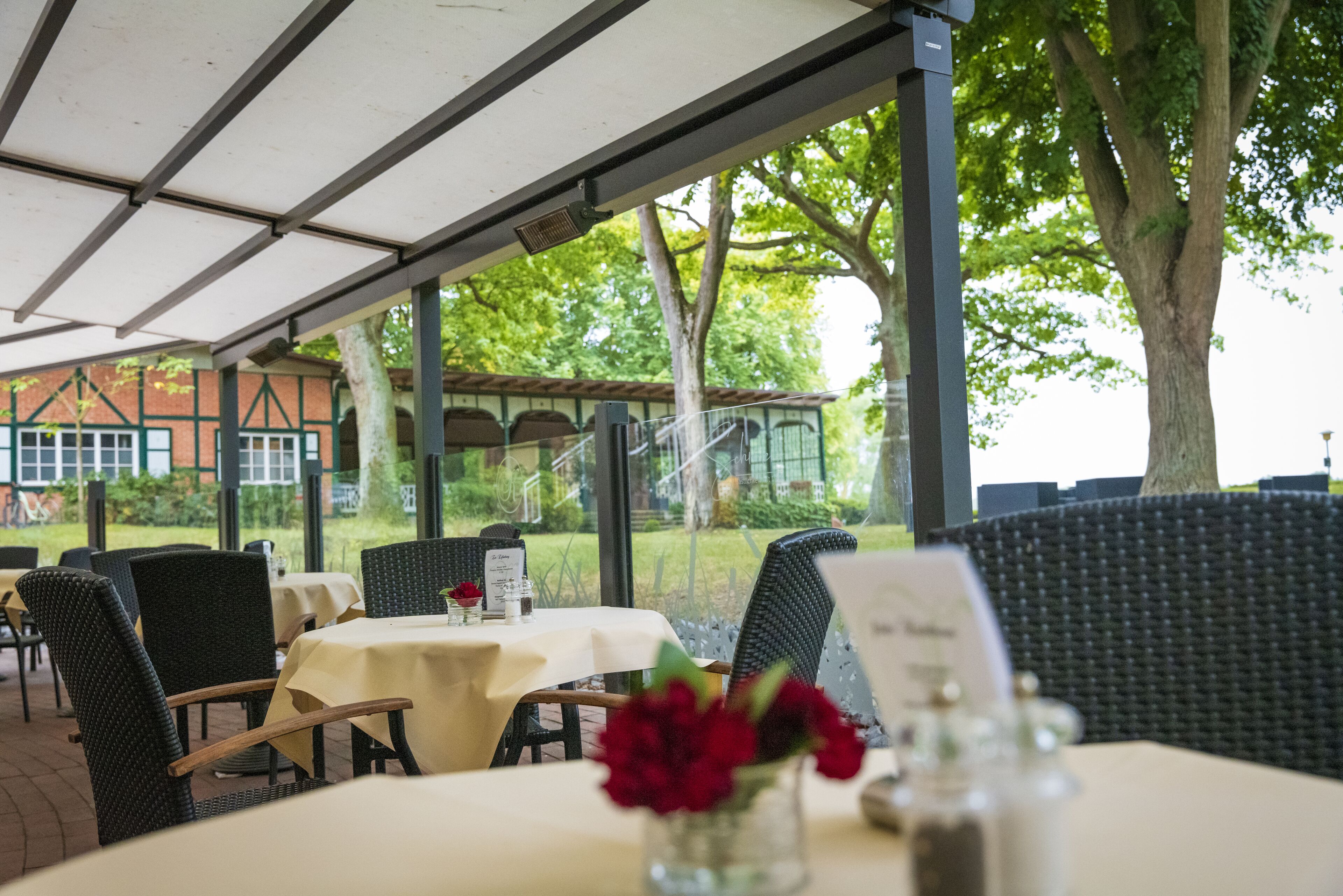 Un área de comedor al aire libre con mesas y sillas, techo cubierto y vista a un edificio y árboles al fondo.