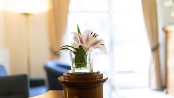 A clear view of a white lily in a glass vase on a wooden pedestal in a well-lit room with a window and a lamp in the background.