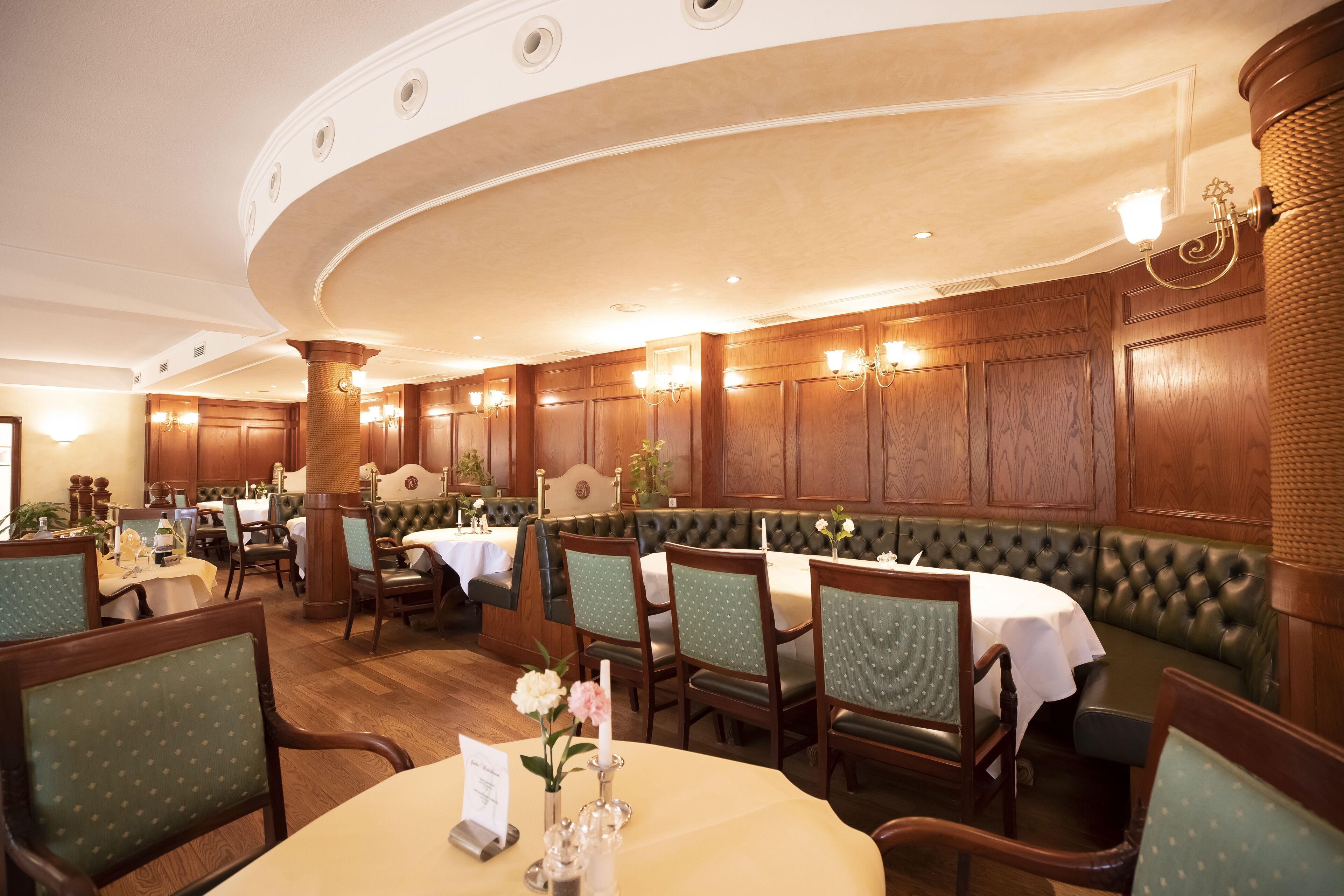 Une salle à manger élégante, avec des tables rondes, des chaises recouvertes d’un tissu vert et des panneaux de bois.