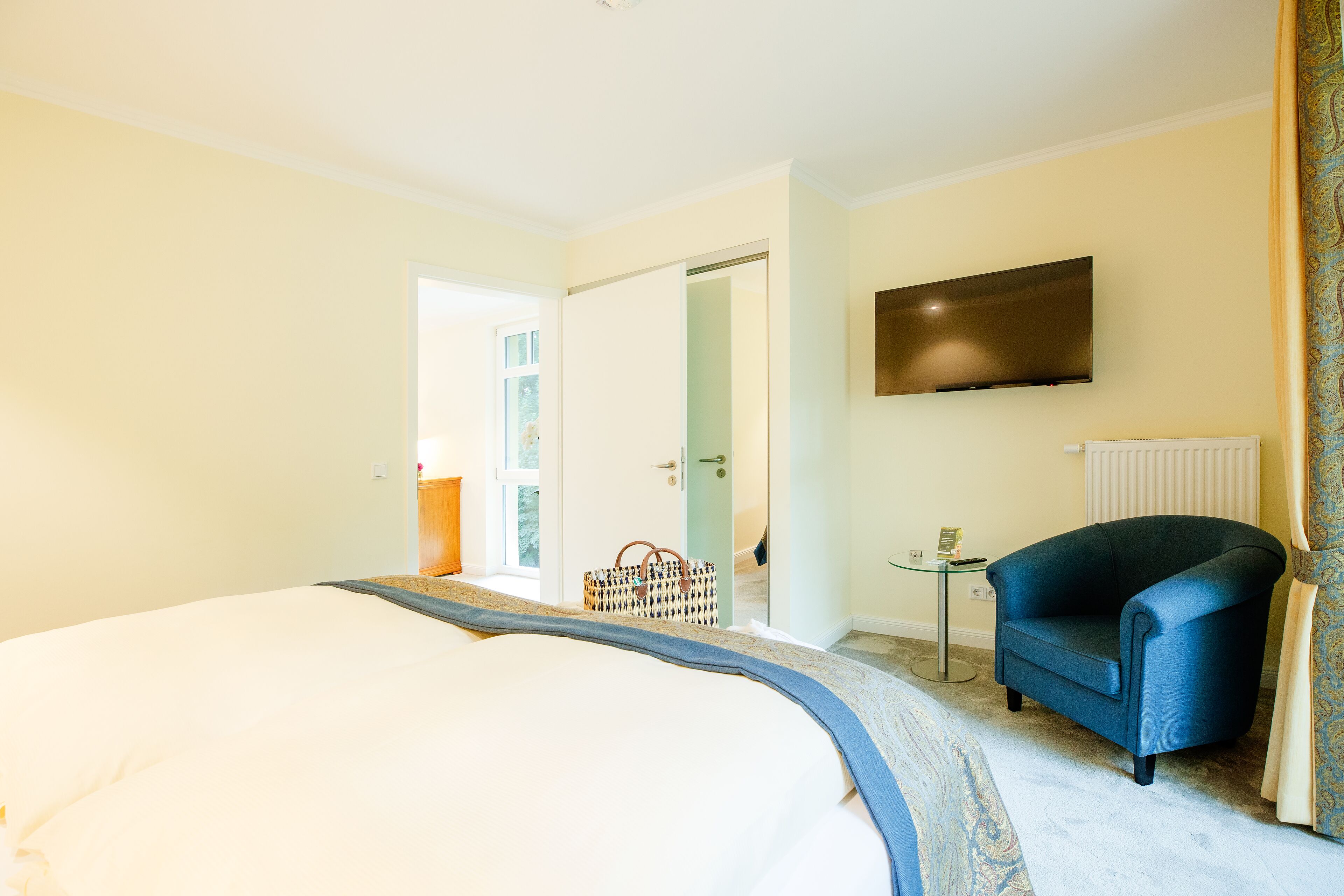 A hotel room with a bed, a blue armchair, a TV mounted on the wall, and a door leading to another room.