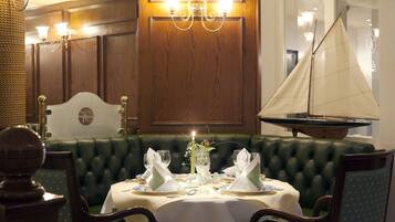 Un comedor con una banca tapizada de verde, una mesa para dos con manteles blancos y un modelo de velero en la pared.