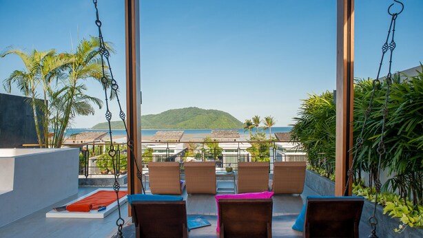 A balcony with wicker chairs, a swing, and a view of the sea and hills. - Almali Luxury Residence (Rawai)