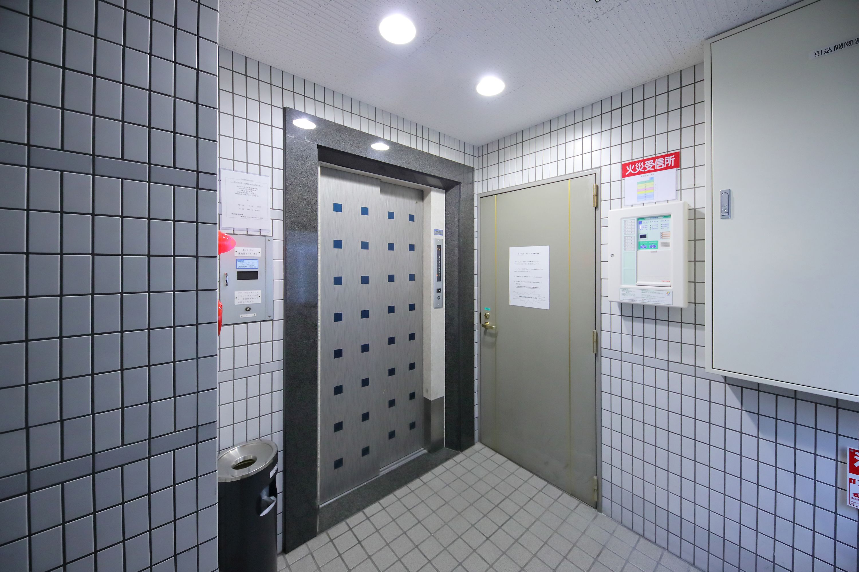 A tiled elevator area with a metallic elevator door, a trash bin, and a fire safety sign.