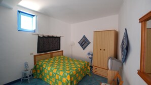 Une chambre avec un lit, une armoire, une petite table, un ventilateur et une fenêtre.
