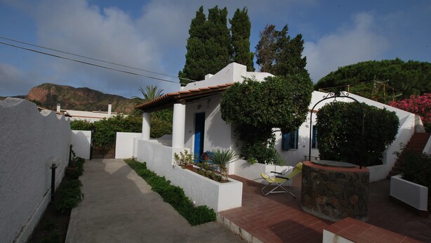 A white building with a blue door, surrounded by greenery and a mountain in the background. - Villino Vulcano (Lipari)