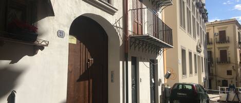 A street view with a building featuring a wooden door, a balcony with metal railings, and a parked car.