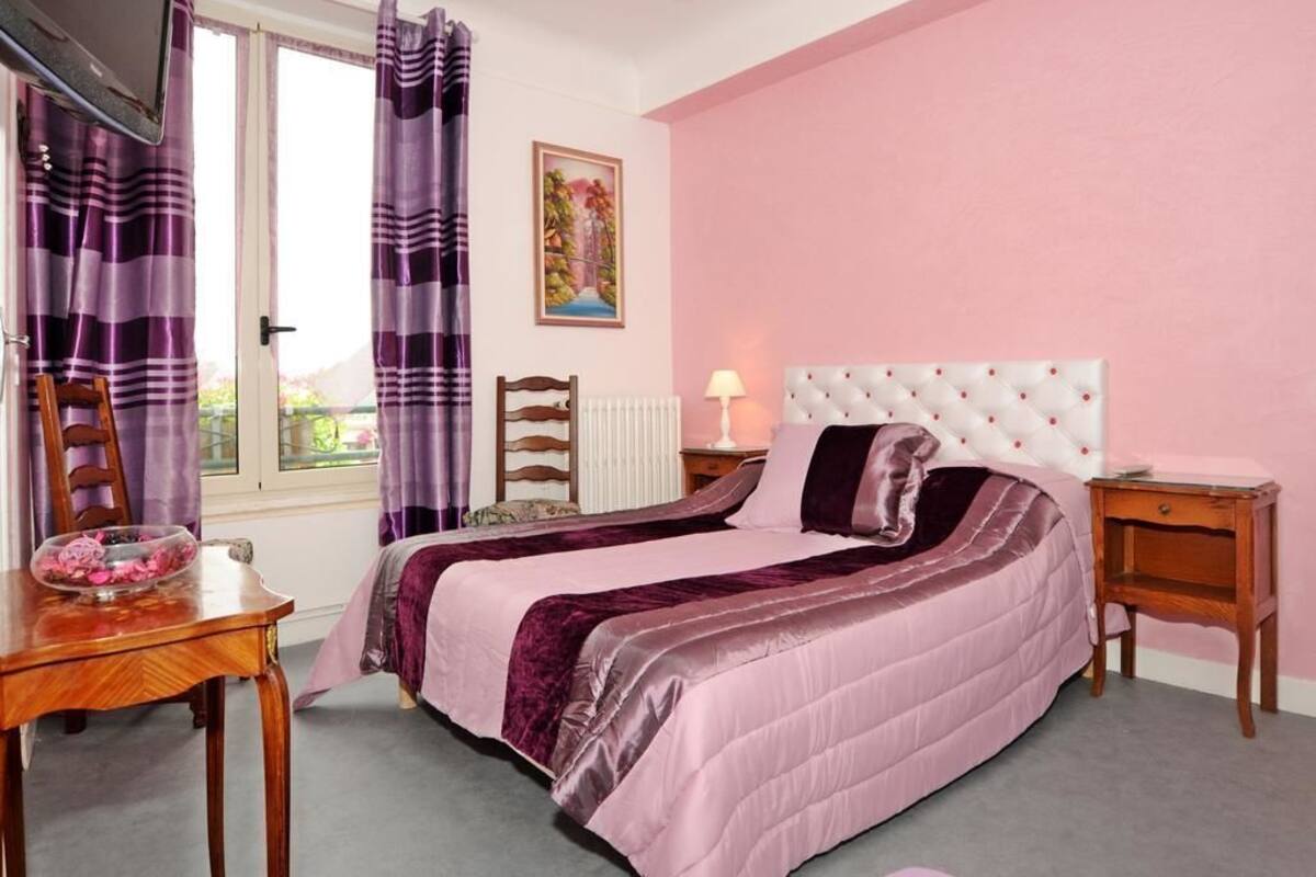 A bedroom with a bed, a wooden chair, a small table with a glass bowl, a nightstand, and a window with curtains.