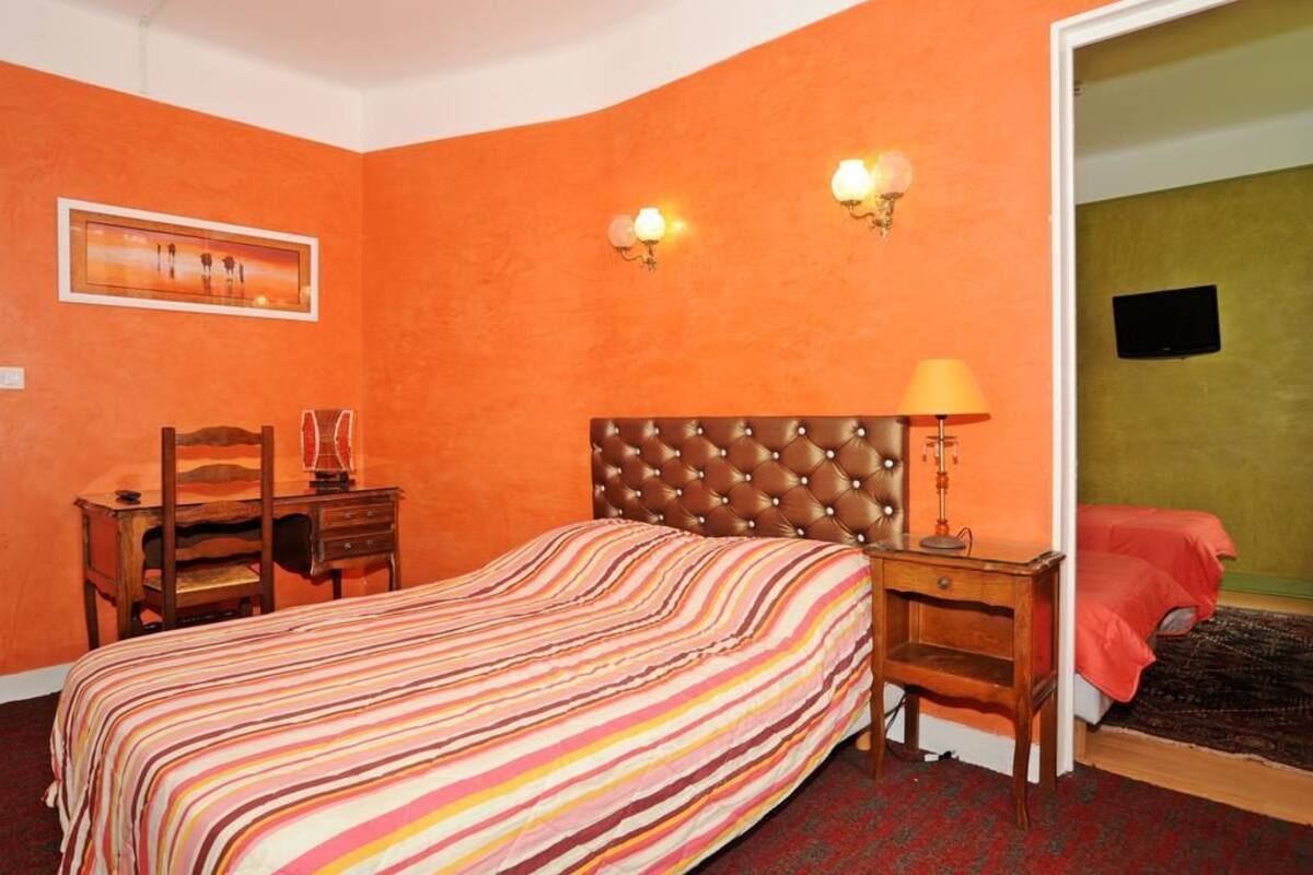 A hotel room with an orange wall, a striped bedspread, a tufted headboard, a desk with a lamp, and a television on the wall.