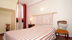 A hotel room with a large bed, a chair, a nightstand, a lamp, a vase, and a window with curtains. - Hôtel la Tour (Sully-sur-Loire)