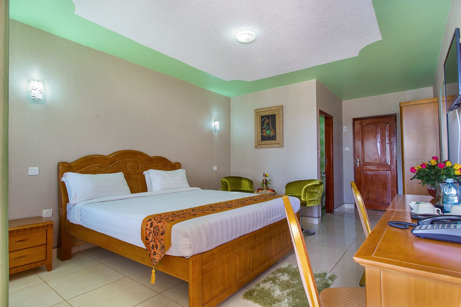 A hotel room with a large bed, a desk with a chair, a wooden door, and a wall-mounted light fixture.