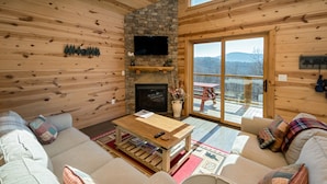 A cozy log cabin interior with a fireplace, wooden walls, a large sofa, and a view through a glass door. - Peaceful Cabin with mountain view! Hot tub, fire-pit and indoor fireplace! (Fairview)