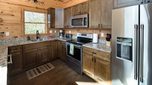 A modern kitchen with wooden cabinets, stainless steel appliances, granite countertops, and a window with a view of trees. - Peaceful Cabin with mountain view! Hot tub, fire-pit and indoor fireplace! (Fairview)
