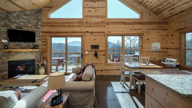 A cozy cabin interior with a stone fireplace, wooden walls, a dining area, and large windows offering mountain views. - Peaceful Cabin with mountain view! Hot tub, fire-pit and indoor fireplace! (Fairview)