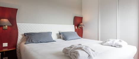 Una cama bien hecha con una colcha blanca, dos almohadas, una toalla blanca enrollada y una mesita de noche con una lámpara.